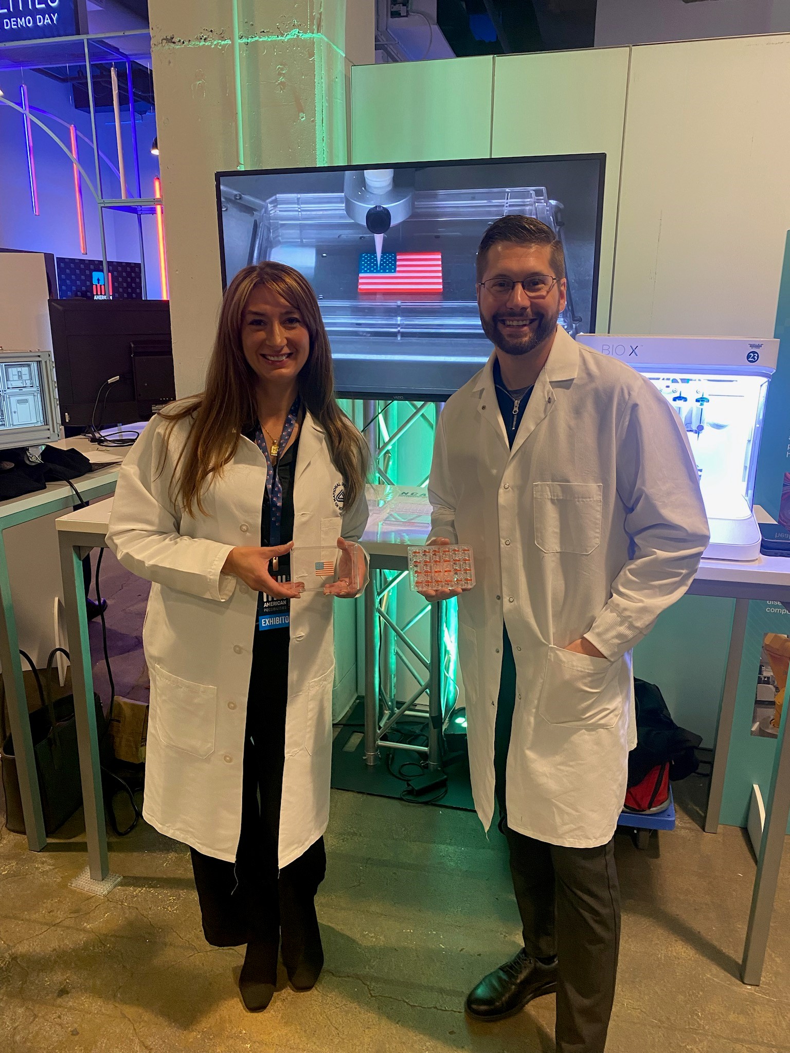 NCATS staff stand in front of lab equipment and a video screen that show how 3-D bioprinting works.